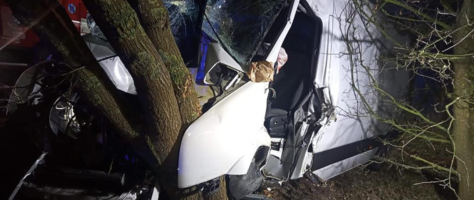 Na zdjęciu biały bus dostawczy po uderzenie w przy drożne drzewo. Jest noc, występuje oświetlenie sztuczne z pojazdów ratowniczych oraz przenośnych najaśnic.