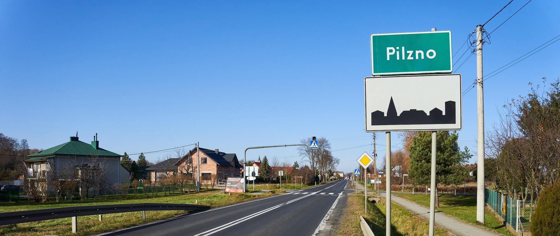 Zdjęcie z poziomu ziemi. Widok na drogę, po jednym pasie w każdym kierunku. Aura jesienna, świeci słońce. W tle widać przejście dla pieszych i znak nad jezdnią informującym o tym. Po prawej stronie widać chodnik, drzewa w kolorach jesieni, domy a przy drodze ustawiony znak z napisem Pilzno. Po lewej stronie drogi widać domy.