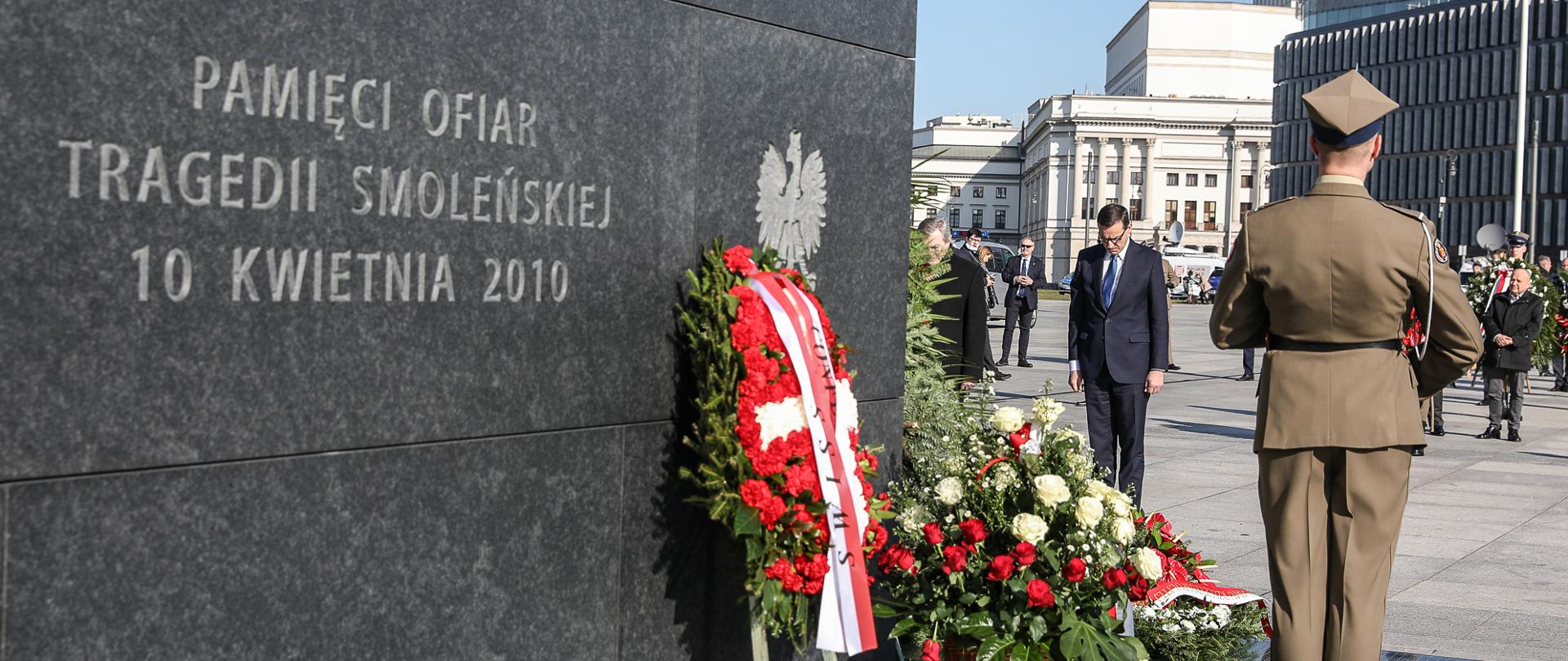 Pomnik Pamięci Ofiar Tragedii Smoleńskiej, pod nim kwiaty. Pomnika pilnuje żołnierz wojska polskiego. Przed pomnikiem hołd ofiarom składa Premier Mateusz Morawiecki.