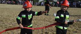 Zawody sportowo-pożarnicze w Jeziorach. Plac ćwiczeń. Trwa jedna z konkurencji zawodów - ćwiczenie bojowe. Końcowy etap ćwiczenia-strącanie przy pomocy strumienia wody, pachołków ustawionych na postumentach.Do wykonania tego zadania jest już gotowych dwóch strażaków - członków MDP. Stojący z przodu trzyma w rękach prądownicę z podłączonym wężem, a jego kolega prawą ręką przytrzymuje wąż, a lewą ma skierowaną do przodu i coś wskazuje. W tle kibice.