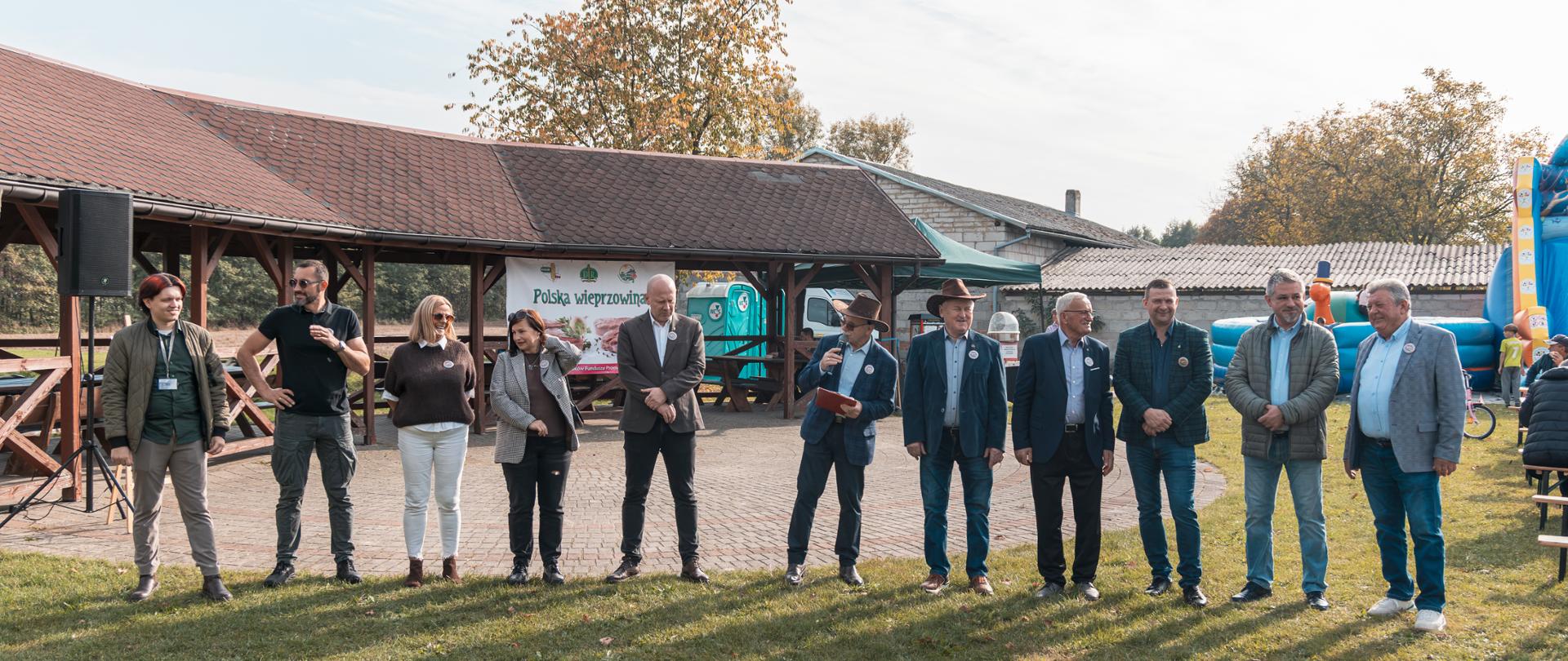 Zdjęcie przedstawia reprezentantów instytucji związanych z rolnictwem oraz władz samorządowych w ilości 11 osób na wykostkowanym placu oraz zielonej trawie przy słonecznej pogodzie i jesiennej atmosferze. W tle jasne niebo oraz drzewa o kolorowo wybarwionych jesiennych liściach.