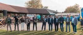 Zdjęcie przedstawia reprezentantów instytucji związanych z rolnictwem oraz władz samorządowych w ilości 11 osób na wykostkowanym placu oraz zielonej trawie przy słonecznej pogodzie i jesiennej atmosferze. W tle jasne niebo oraz drzewa o kolorowo wybarwionych jesiennych liściach.
