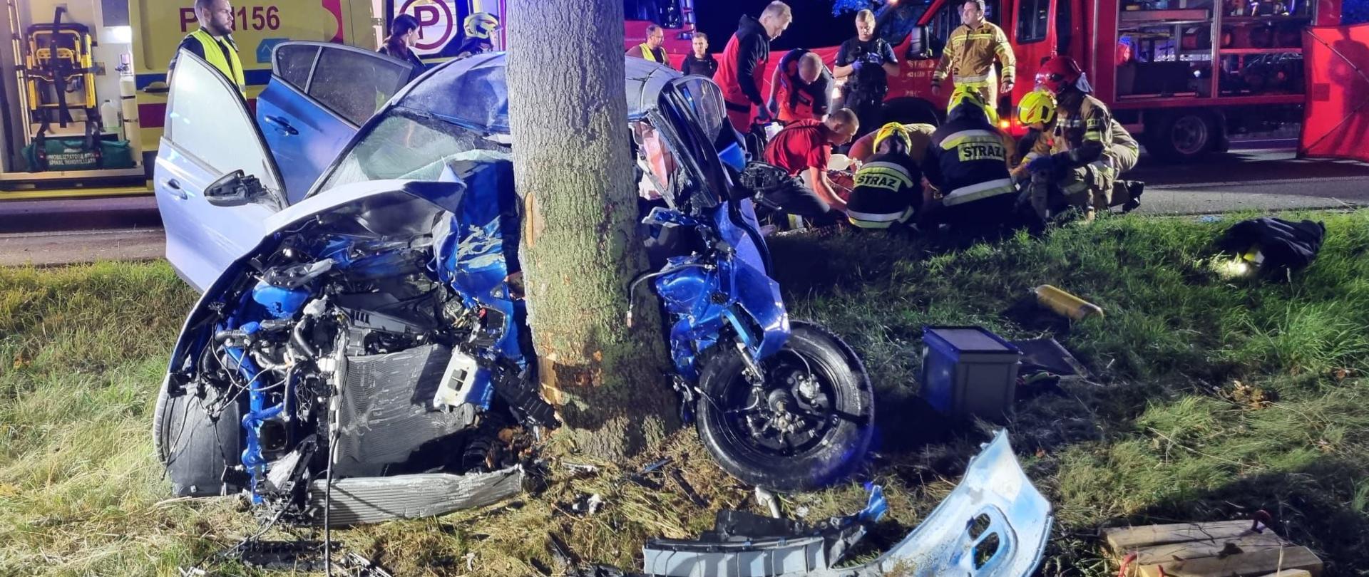 Rozbity na drzewie samochód osobowy, w tle strażacy i ratownicy ZRM udzielający poszkodowanemu pomocy medycznej, dalej karetka ZRM i pojazdy strażackie.