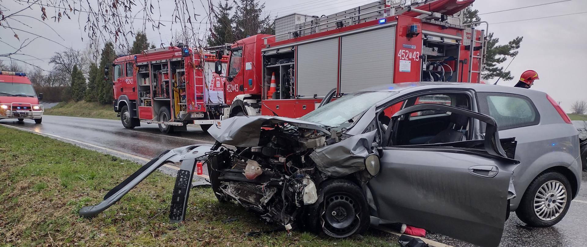 Na pierwszym planie rozbity samochód marki Fiat Punto. W drugim planie widać 3 samochody Państwowej Straży Pożarnej oraz strażaka zabezpieczającego teren działań.