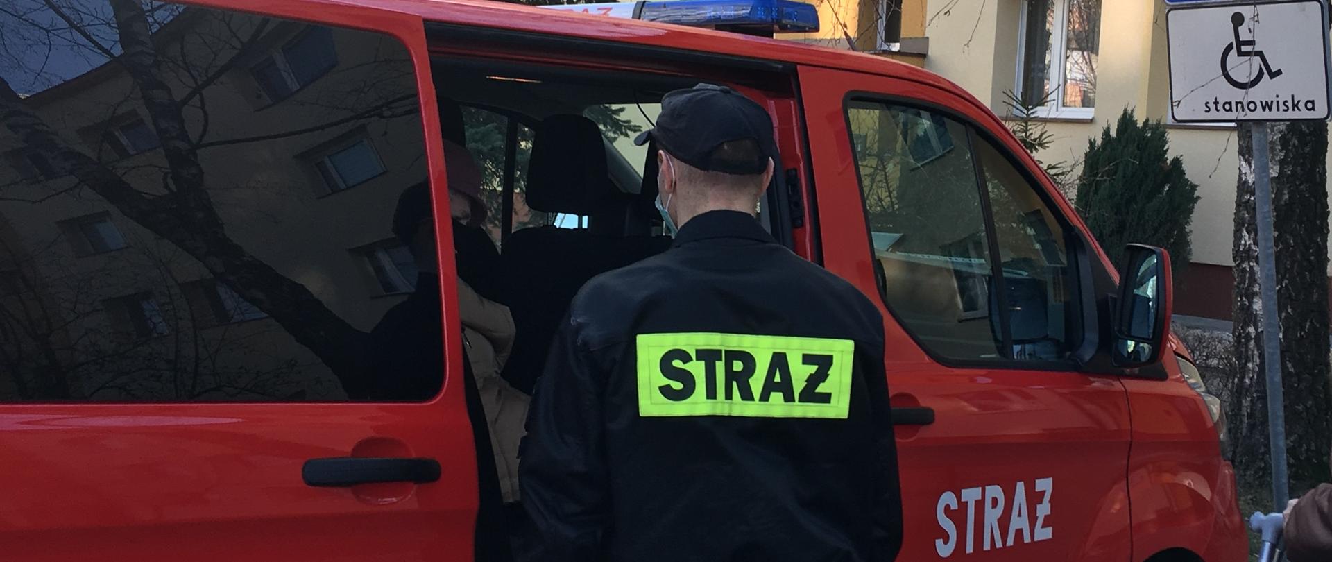 Na zdjęciu widać strażaka, który stoi przed zaparkowanym na parkingu osiedlowym samochodem pożarniczym. W samochodzie siedzi osoba, która zostanie zawieziona do przychodni na szczepienie.