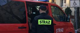 Na zdjęciu widać strażaka, który stoi przed zaparkowanym na parkingu osiedlowym samochodem pożarniczym. W samochodzie siedzi osoba, która zostanie zawieziona do przychodni na szczepienie. W tle widać blok mieszkalny.