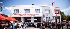 Zdjęcie grupowe uczestników uroczystości na placu przed remizą Ochotniczej Straży Pożarnej w Pionkach. Po lewej stronie stoją dwa czerwone namioty. Po prawej stronie stoi maszt z biało-czerwoną flagą. Nad budynkiem widoczne jest pogodne błękitne niebo z małymi białymi chmurami. 