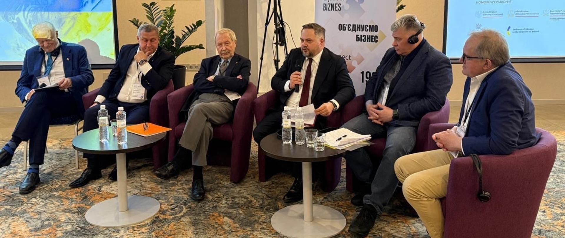 Wiceminister Konrad Gołota i inni paneliści siedzą na fotelach. W tle ekrany i roll up.