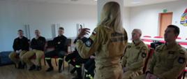 Świetlica komendy. Na krzesłach siedzą strażacy uczestniczący w zajęciach. Przed nimi stoi pani psycholog, która prowadzi psychoedukację. Wszyscy strażacy są umundurowani. W tle na ścianie widoczne jest logo PSP i drzwi ewakuacyjne. Z lewej strony, przy ścianie ustawione są stoły.