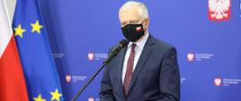 Wicepremier i minister rozwoju, pracy i technologii Jarosław Gowin w maseczce na twarzy stojący przed mównicą, po lewej stronie wicepremiera flagi Polski i UE