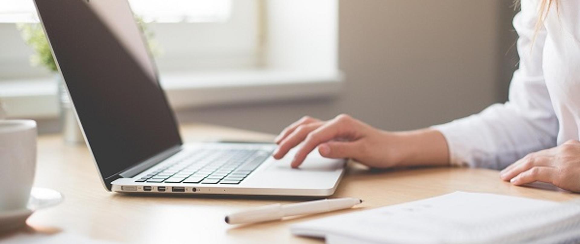 kobieta pracująca przy laptopie. Na biurku widać też dokumenty i długopis