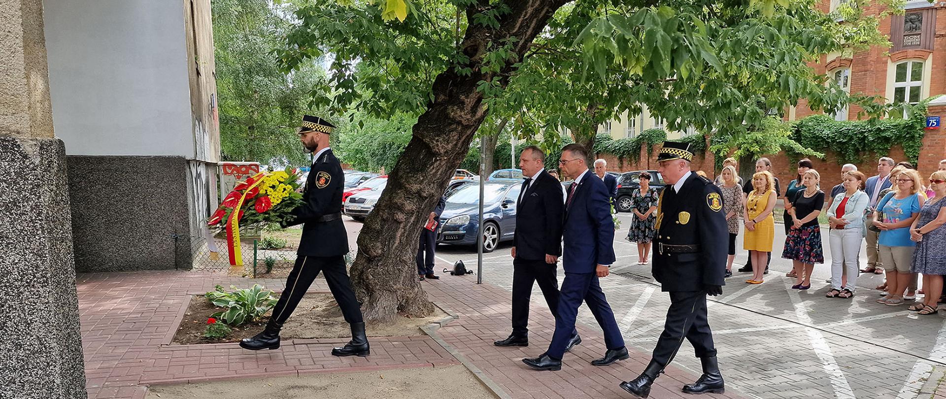 Delegacja mężczyzn podczas marszu z wieńcem w stronę tablicy pamiątkowej. W tle pracownicy Inspekcji Sanitarnej.