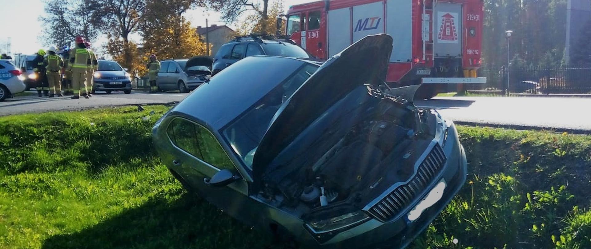 Zdjęcie przedstawia jeden z uszkodzonych w kolizji samochodów osobowych, który znalazł się w przydrożnym rowie. Za nim, na jezdni stoi samochód pożarniczy, z lewej widać fragment radiowozu oraz przemieszczających się po drodze strażaków.
