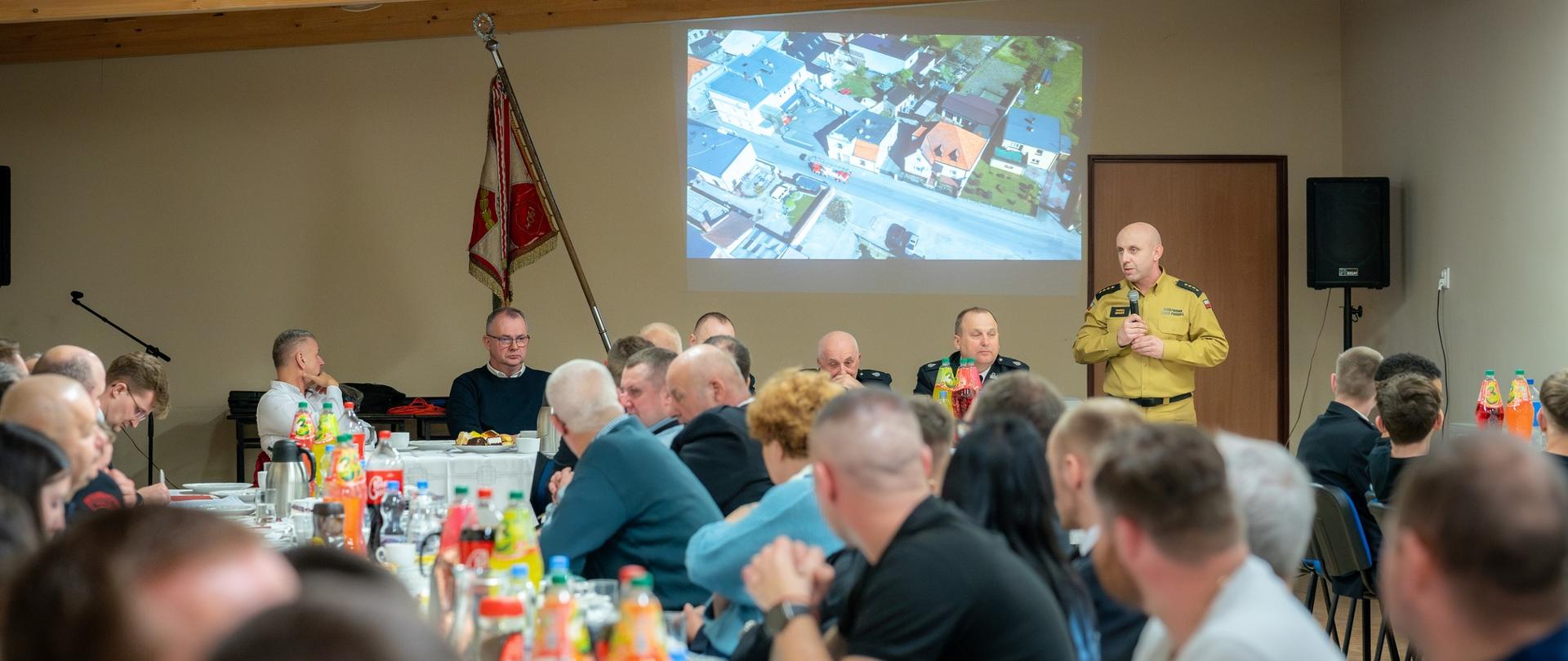 Grupa ludzi słuchająca wypowiedzi strażaka. W tle sztandar jednostki OSP.