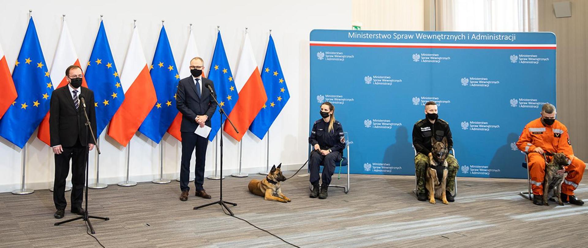 Na zdjęciu z prawej strony widać na tle flag polskich i unijnych stojących przy mikrofonach na statywach Ministra i Wiceministra MSWiA ubranych w ciemne garnitury. z prawej strony widać na tle niebieskiej planszy z napisami Ministerstwo Spraw Wewnętrznych i Administaracji siedzących na krzesełkach funkcjonariuszy policji, straży granicznej oraz straży pożarnej. Funkcjonariusze ubrani są w umundurowanie służbowe. Przy każdym z funkcjonariuszy siedzi pies.