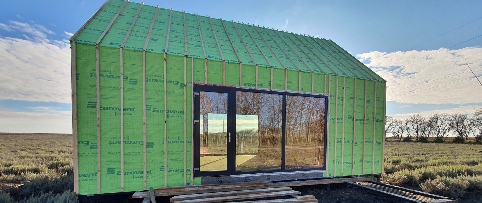 Świeżo wybudowany, nieduży budynek stojący na środku rozświetlonej słońcem łąki.