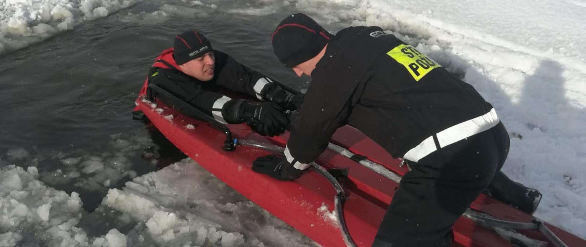 Na zdjęciu mamy 2 strażaków w skafandrach suchych. Jeden jest w przeręblu a drugi na saniach lodowych.Akwen jest pokryty lodem i śniegiem.
