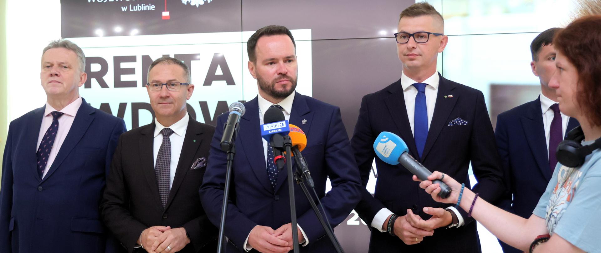 ojewoda Lubelski Krzysztof Komorski (w centrum, w ciemnym garniturze) wraz z wicewojewodą Andrzejem Majem (drugi od lewej), dyrektorem Oddziału ZUS w Lublinie Piotrem Waszakiem (pierwszy z lewej) oraz dyrektorem Oddziału Regionalnego KRUS w Lublinie Mateuszem Winiarskim (pierwszy z prawej), stoją przed mikrofonami z logotypami TVP3 Lublin i Polsat News, podczas konferencji prasowej w Lubelskim Urzędzie Wojewódzkim. Za nimi widoczny jest duży ekran z napisem "LUBELSKI URZĄD WOJEWÓDZKI w Lublinie" oraz częściowo widocznym napisem "RENTA WDOWIA", co wskazuje na temat spotkania – nowy program rządowy.