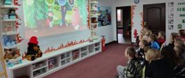 Dzieci ze Szkoły Podstawowej nr 3 w Łowiczu siedzą na krzesełkach i oglądają film wyświetlany na ścianie w tle czerwona hydronetka oraz dwoje drzwi prowadzących do kolejnych pomieszczeń na ścianie kolorowe infografiki na podłodze ciemnobrązowa wykładzina 