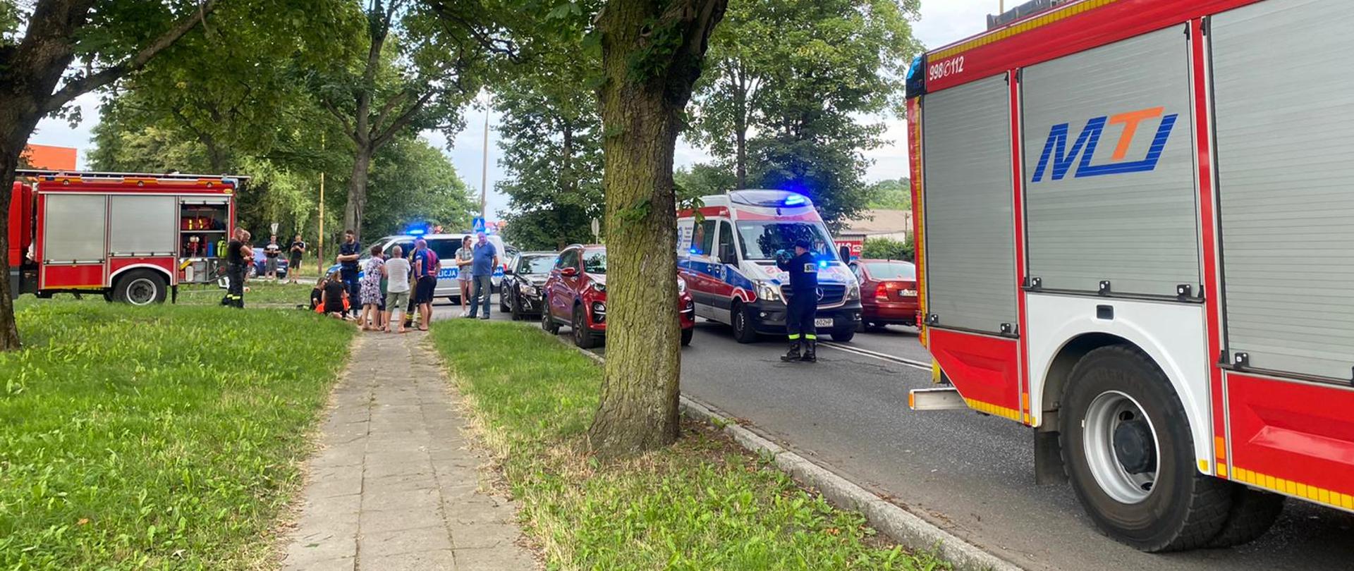 poszkodowani stoją na chodniku przy pojazdach, które uległy kolizji, za nimi w tle radiowóz Policji, drogi zablokowane przez pojazdy pożarnicze