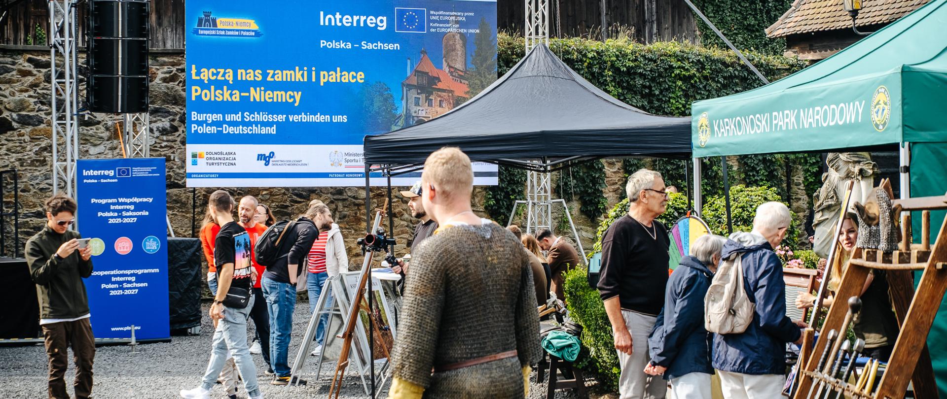 Grupa osób w plenerze przechodzi obok stoisk 