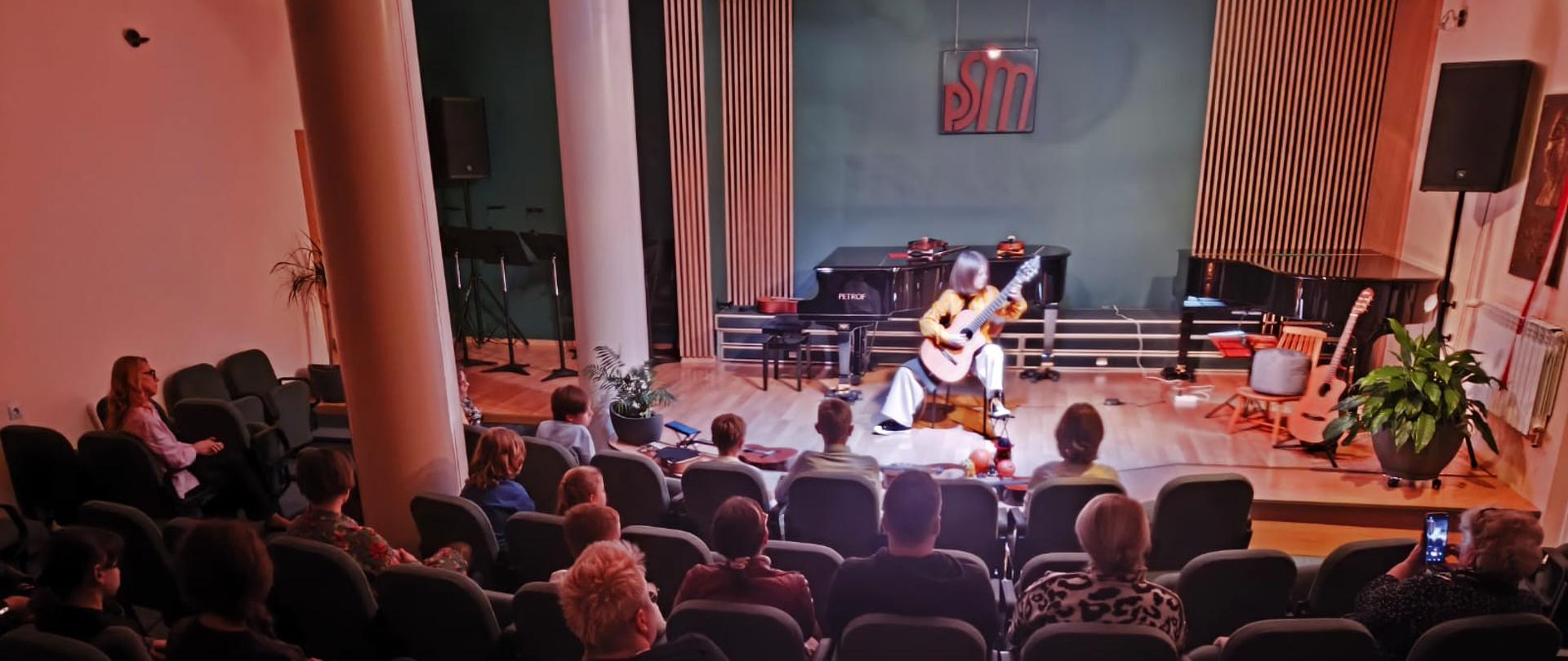 Widownia siedzi w rzędach krzeseł w niewielkiej sali koncertowej, oglądając występ muzyczny. Na scenie artysta gra na gitarze klasycznej, siedząc na krześle. W tle widoczne są fortepian oraz logo z literami 'PSM' na ścianie. Sala jest przytulna, z drewnianą podłogą i roślinami doniczkowymi, które dodają ciepłego charakteru wnętrzu