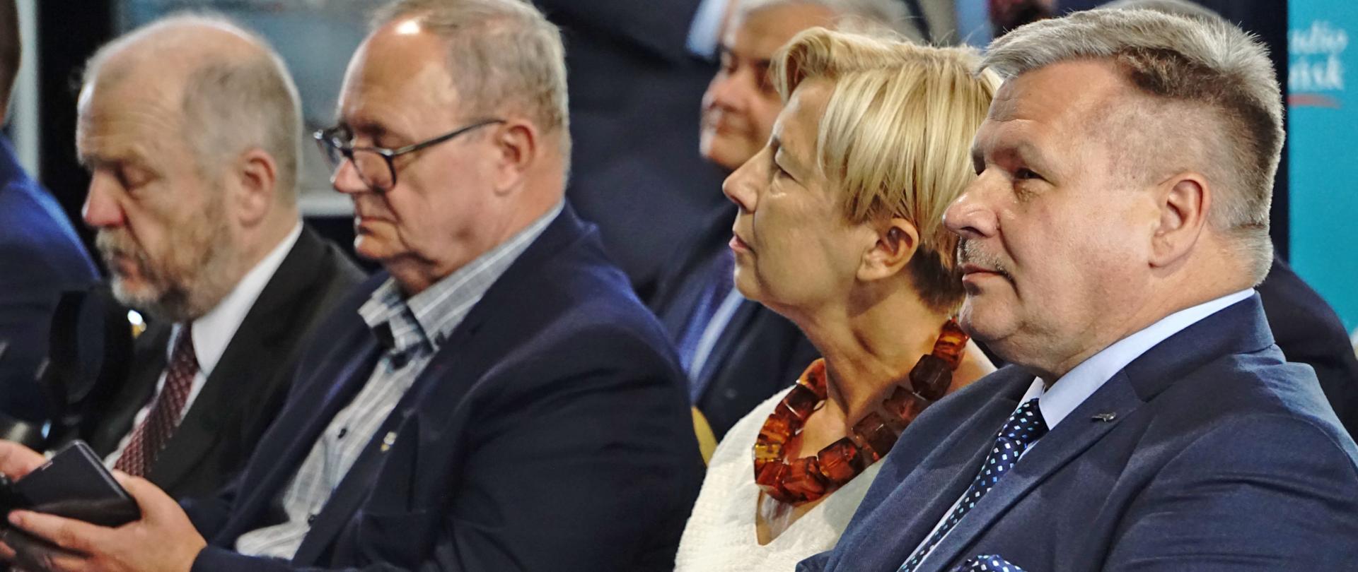 Grupa osób siedzi na krzesłach, ich wzrok jest skierowany w jedną stroną 