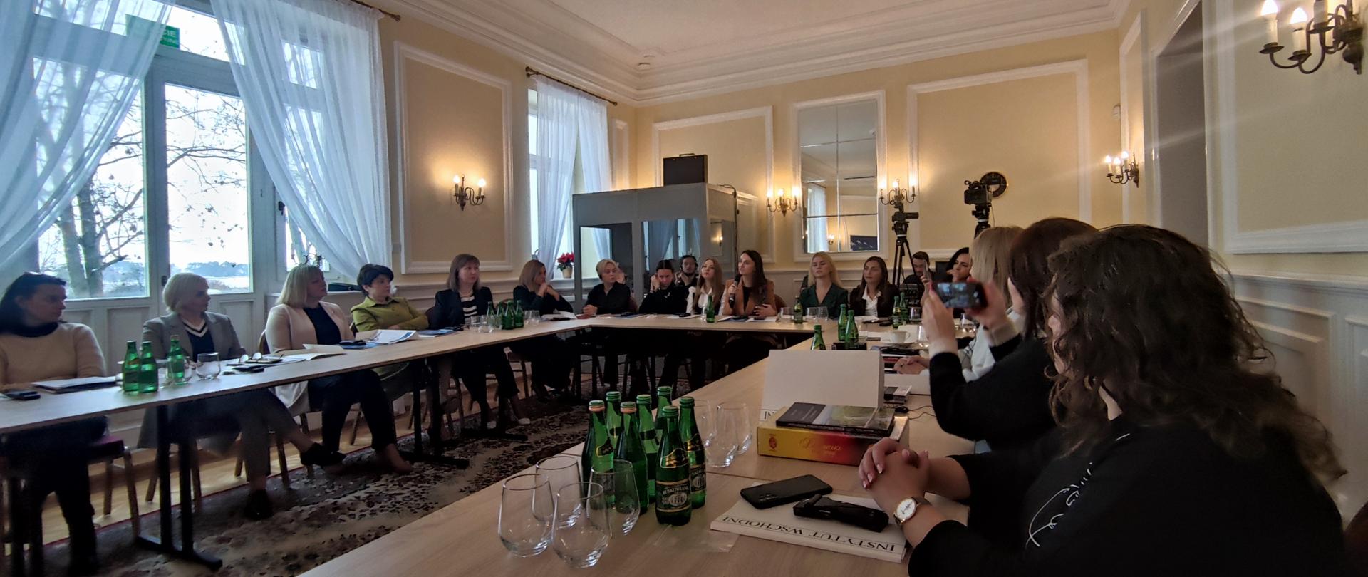 W sali konferencyjnej przy ustawionych w prostokąt stołach siedzą uczestniczki warsztatów i słuchają przemawiającej kobiety