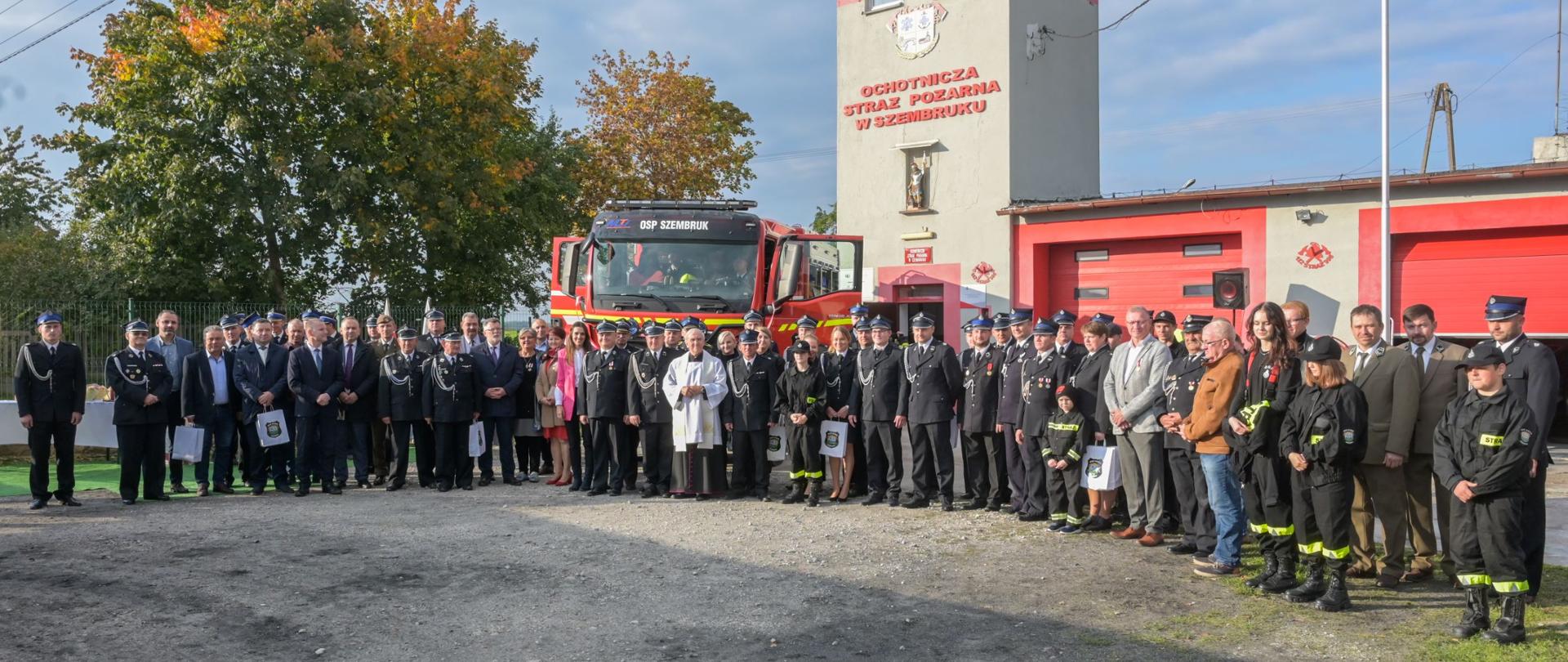 Zdjęcie przedstawia czerwono-biały ciężarowy pojazd gaśniczy. Przed nim w łuku stoi kilkadziesiąt osób (strażacy i osoby cywilne) pozując do zdjęcia. W tle budynek z czerwonymi bramami wyjazdowymi i wieżą oraz zielone drzewa.