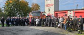 Zdjęcie przedstawia czerwono-biały ciężarowy pojazd gaśniczy. Przed nim w łuku stoi kilkadziesiąt osób (strażacy i osoby cywilne) pozując do zdjęcia. W tle budynek z czerwonymi bramami wyjazdowymi i wieżą oraz zielone drzewa.