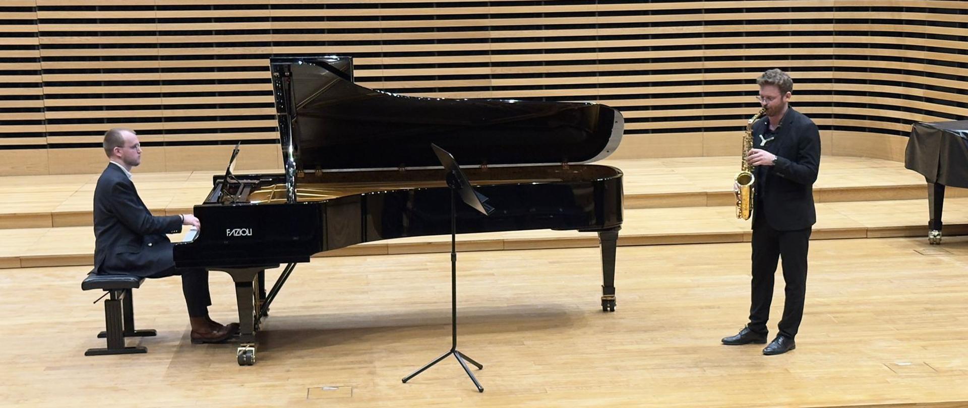 Mężczyzna gra na saksofonie, drugi mężczyzna akompaniuje mu na fortepianie.