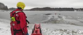 Na ośnieżonym brzegu jeziora strażak obserwuje akcje ratowniczą wyciągania mężczyzny z przerębli.