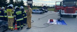 na zdjęciu strażacy w trakcie wykonywania dostępu do osoby poszkodowanej
w trakcie założenia egzaminacyjnego, w tle pojazd pożarniczy