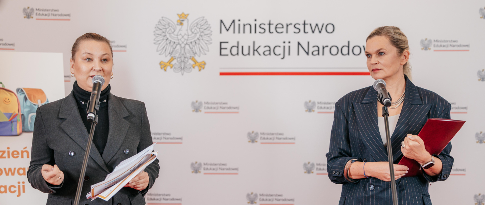 Dwie kobiety stojące na tle ściany z logotypem Ministerstwa Edukacji Narodowej i powtarzającym się napisem "Ministerstwo Edukacji Narodowej". Kobieta po lewej w ciemnym żakiecie i golfie mówi do mikrofonu, trzymając dokumenty. Kobieta po prawej w granatowym garniturze w paski trzyma czerwoną teczkę i również stoi przy mikrofonie.