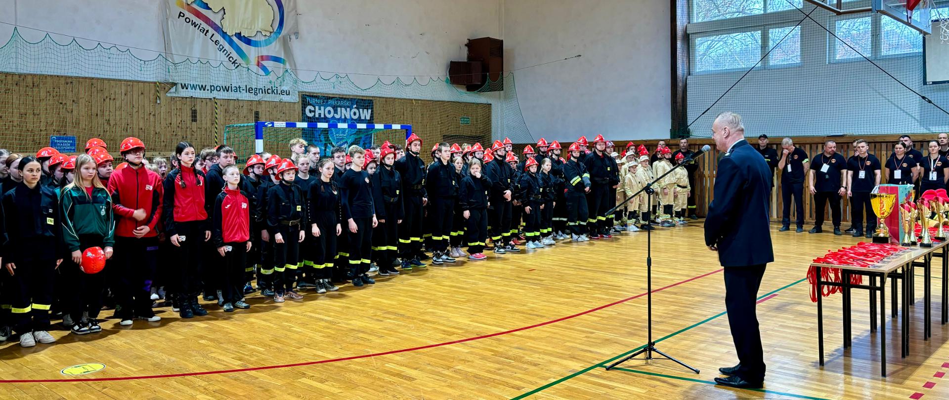 dzieci w mundurach strażackich stoją na zbiórce w dwuszeregu, do nich przemawia druh prezes OSP