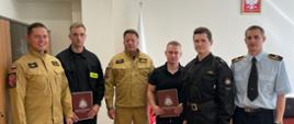 Na zdjęciu widoczne jest 5 osób, od lewej: Zastępca Komendanta, nowo przyjęty strażak, Komendant Powiatowy, nowo przyjęty pracownik cywilny, Zastępca dowódcy JRG, Funkcjonariusz z sekcji organizacyjno-kadrowej KP PSP w Kętrzynie. W tle flaga oraz wiszący na ścianie herb Polski.