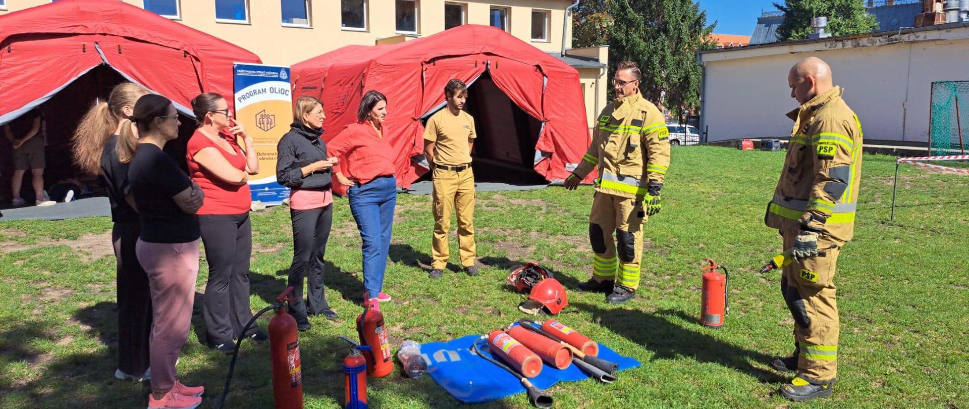 Pokaz sprzętu gaśniczego na trawie przed czerwonym namiotem pneumatycznym. Dwóch strażaków w piaskowych mundurach bojowych stoi przy niebieskiej plandece, na której ułożone są węże, prądownica i gaśnice. Prezentują sprzęt grupie pięciu kobiet cywilów, które uważnie słuchają i obserwują. W tle widać budynek z czerwoną dachówką i inne czerwone namioty.