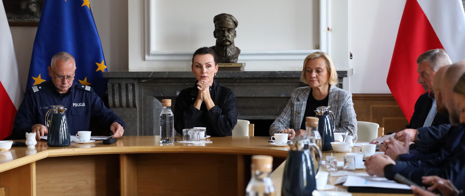 Na zdjęciu widoczna jest narada, w której uczestniczy grupa osób siedzących przy dużym, jasnym, drewnianym stole. W centralnej części kadru, naprzeciwko obiektywu, zasiada kilka głównych postaci. Po lewej stronie stołu siedzi mężczyzna w granatowym mundurze z napisem „POLICJA”. Obok niego, z dłońmi splecionymi przed sobą, siedzi kobieta ubrana w czarny strój, a po jej prawej stronie znajduje się kolejna kobieta w jasnej, wzorzystej marynarce. Wzdłuż prawego skrzydła stołu rzędem siedzą pozostali uczestnicy narady, widoczni głównie profilu lub od tyłu. Na stole stoją dzbanki z napojami, karafki z wodą, filiżanki. W tle, za uczestnikami spotkania, widoczny jest kominek, na którym stoi popiersie oraz zawieszone są flagi Polski i Unii Europejskiej.