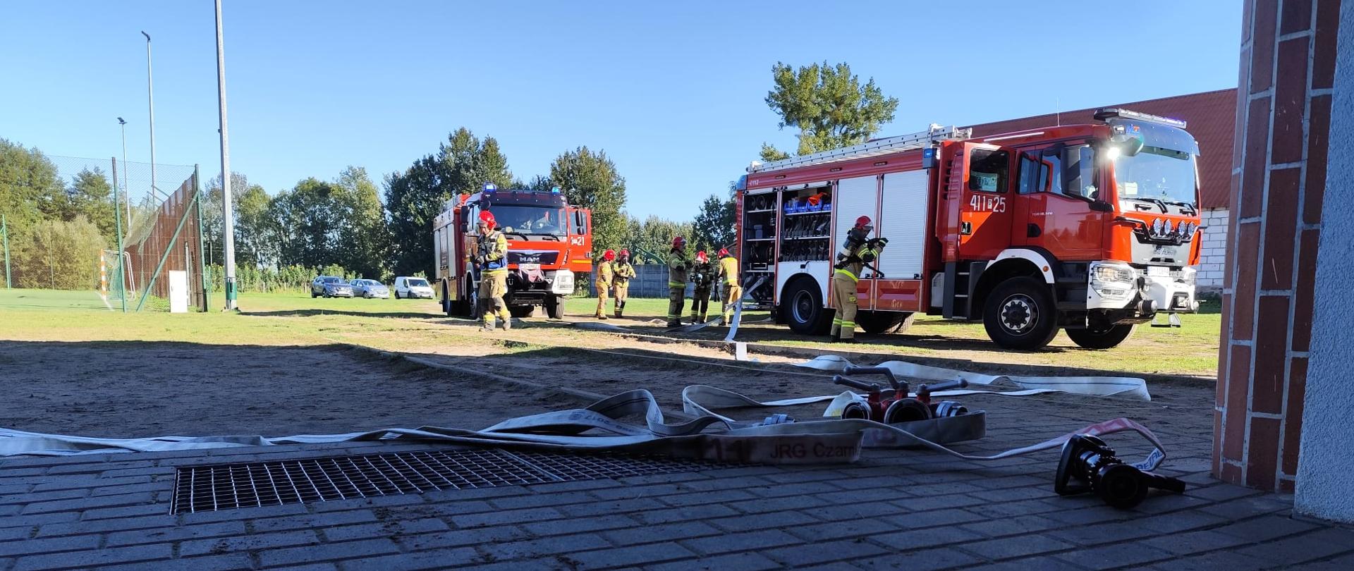 Wozy strażackie ustawione na terenie szkoły, strażacy przygotowują sprzęt do ćwiczeń, widoczne rozwinięte węże pożarnicze.