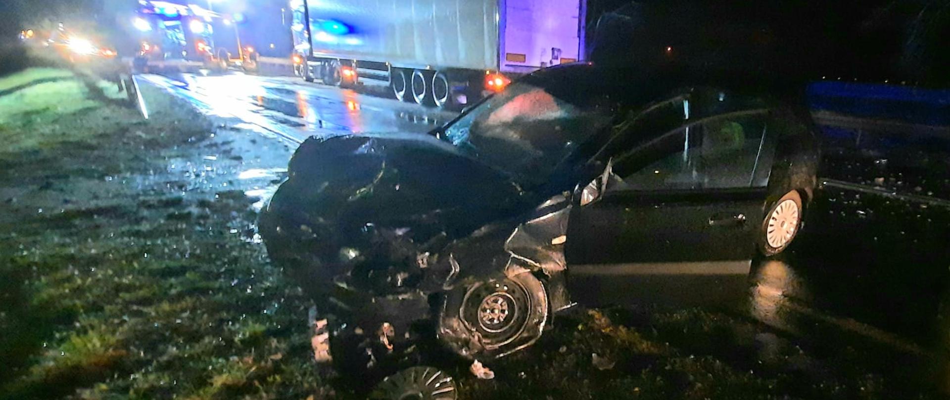 Samochód osobowy stoi w poprzek drogi. Na drugim planie strażacy działają przy zdarzeniu.