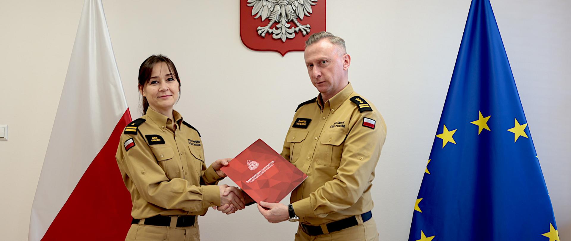 Na zdjęciu widać dwoje funkcjonariuszy Państwowej Straży Pożarnej stojących naprzeciw siebie w gabinecie. Kobieta znajduje się po lewej stronie, mężczyzna po prawej. Mężczyzna to starszy brygadier Damian Legierski śląski komendant wojewódzki. Oboje ubrani są w jednakowe, piaskowe mundury służbowe z oznaczeniami PSP oraz naszywkami z flagą Polski na rękawach. Obie osoby patrzą w kierunku aparatu. Między nimi trwa uścisk dłoni. Drugą ręką wspólnie trzymają czerwoną teczkę z dokumentem, aktem powierzenia pełnienia obowiązków zastępcy naczelnika wydziału kadr.W tle, na ścianie, znajduje się godło Polski – biały orzeł na czerwonym tle. Po lewej stronie stoi flaga Polski (biało-czerwona), a po prawej flaga Unii Europejskiej (niebieska z żółtymi gwiazdami). 