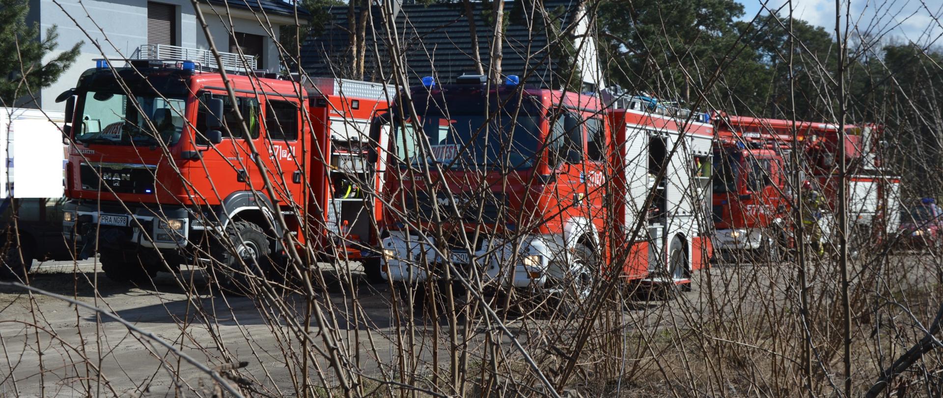 Pożar budynku mieszkalnego w Sierakowie. Na drodze gruntowej - na tle budynków mieszkalnych stoją cztery wozy straży pożarnej. Obok samochodu z podnośnikiem hydraulicznym widoczna jest sylwetka strażaka w czerwonym hełmie. Na pierwszym planie krzewy.