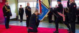 Zdjęcie dokumentuje jeden z najbardziej podniosłych momentów w ceremoniale strażackim – pożegnanie ze sztandarem odchodzącego Komendanta Powiatowego Państwowej Straży Pożarnej w Wołowie. Na środku czerwonego dywanu klęczy na jedno kolano funkcjonariusz w ciemnym mundurze galowym. Z szacunkiem pochyla się nad sztandarem jednostki, chwytając oburącz za dół jego płata, aby symbolicznie go ucałować. Sztandar: Jest to niebieski płat z haftowanym napisem „Komenda Powiatowa Państwowej Straży Pożarnej w Wołowie” oraz widoczną datą „2025”. Drzewce zwieńczone jest metalową głowicą w kształcie orła, a do płata przywiązana jest biało-czerwona wstęga. Po prawej stronie funkcjonariusz z pocztu sztandarowego, w czarnych rękawiczkach i z biało-czerwoną szarfą, pochyla sztandar w stronę klęczącego komendanta. Obok niego inny strażak stoi w postawie zasadniczej, oddając honor poprzez salutowanie. W tle, po lewej stronie i wzdłuż bram garażowych, stoją inni oficerowie oraz strażacy w pełnym umundurowaniu wyjściowym. Zachowują nieruchomą, uroczystą postawę, obserwując ceremonię. Uroczystość odbywa się wewnątrz hali garażowej komendy. W tle widać białe bramy segmentowe, rury wyciągowe do spalin oraz fragmenty ścian w kolorze oliwkowym i bordowym.