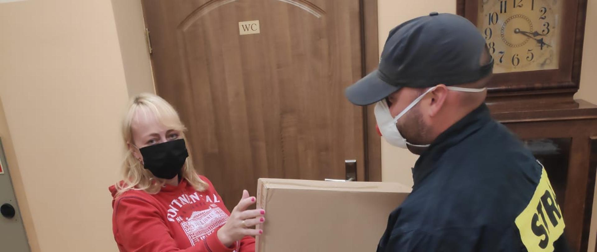 Zdjęcie przedstawia strażaka przekazującego karton ze środkami ochrony indywidualnej pracownicy jednej z placówek oświatowych na tle drzwi i zegara. 