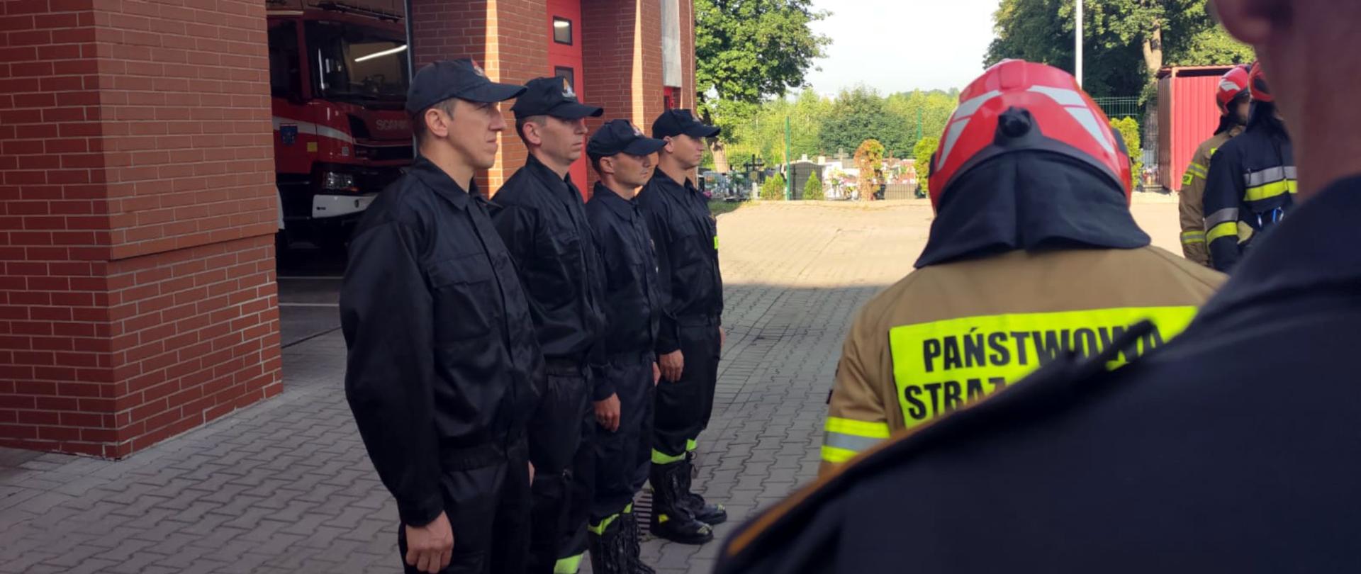 Zdjęcie przedstawia czterech strażaków w umundurowaniu koszarowym. W tle otwarty garaż w obiekcie JRG.