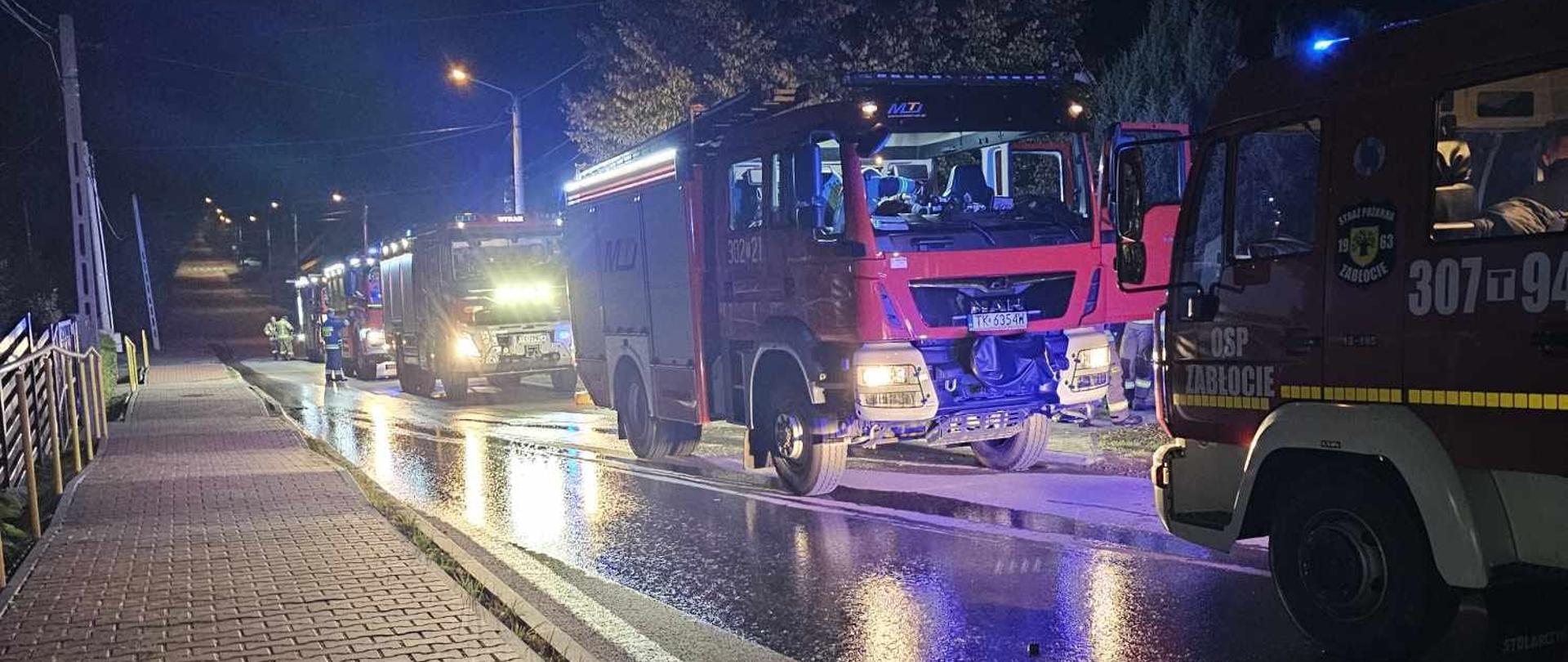 Zdjęcie przedstawia sznur samochodów pożarniczych, które przybyły na ratunek mieszkańcom domu jednorodzinnego w Jaworzu. Pojazdy stoją w szeregu na mokrej jezdni.
