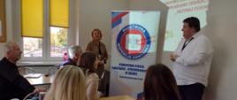 sala wykładowa, pięć osób siedzących przy stolikach, dwie osoby stojące, w tle baner Państwowej Inspekcji Sanitarnej w Śremie