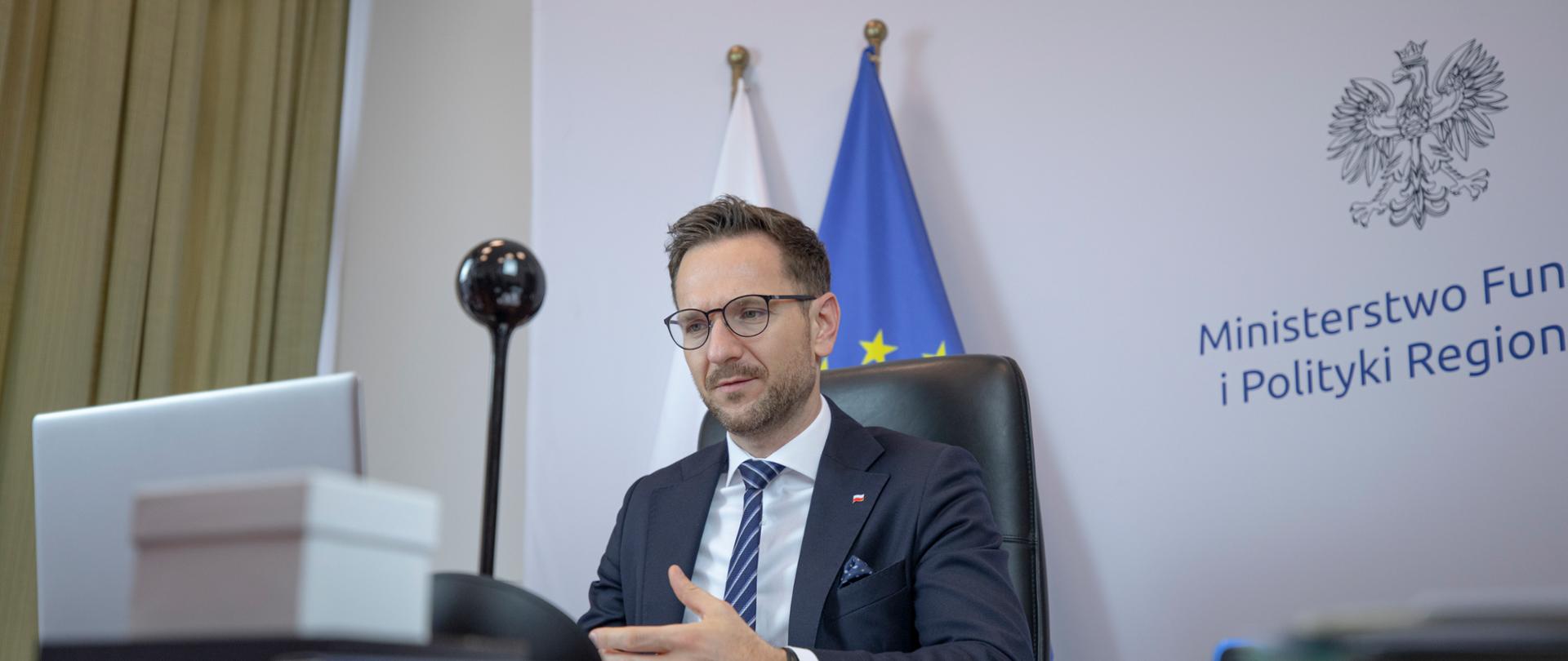 Wiceminister Waldemar Buda siedzi w gabinecie przed monitorem. Na blacie lampka i pudełko. Za ministrem ścianka ministerstwa i dwie flagi PL i UE.