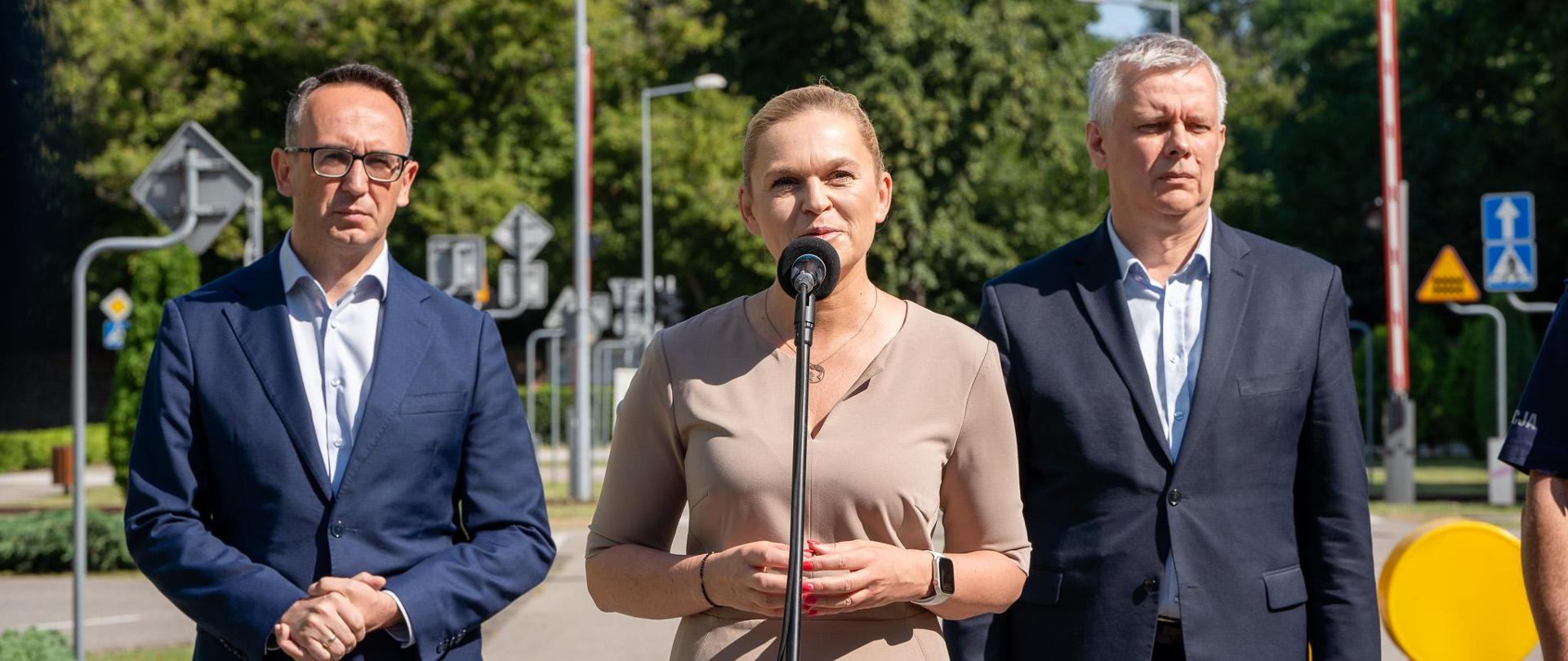 Minister Edukacji Barbara Nowacka stoi przed mikrofonem ustawionym na statywie i mówi. Za nią stoją Minister Infrastruktury Dariusz Klimczak oraz szef MSWiA Tomasz Siemoniak. W tle miasteczko ruchu drogowego. 