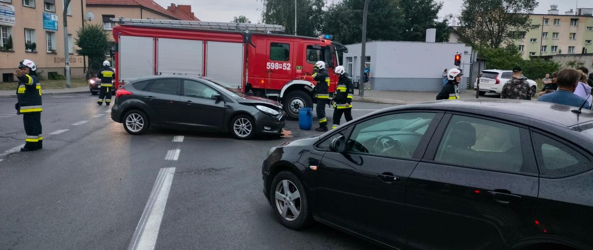 Widać dwa uszkodzone samochody na skrzyżowaniu, kilku strażaków oraz w tle samochód straży pożarnej